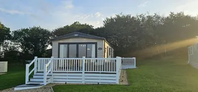 a small cabin with a white fence in the grass at Hot Tub Lodge Cornwall - Meadow Lakes Holiday Park in St Austell