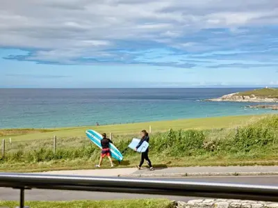 two people walking down a sidewalk with surfboards and the ocean at 15 Ocean Gate Newquay in Crantock
