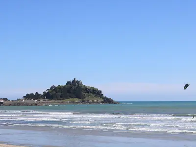 a kite flying over a beach with an island in the ocean at Little Wren Cottage in Marazion