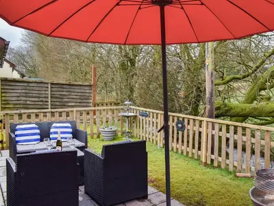 a red umbrella on a patio with two chairs and a table at 15 Pebble Cottage in Camelford