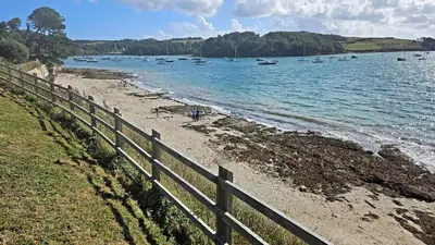 a fence next to a beach with boats in the water at The Red House Annexe in Saint Mawes
