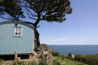 a blue building with a tree next to the ocean at Shepherd's Hut Gorran Haven Panoramic Ocean Views in Gorran Haven