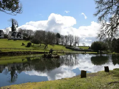 a pond in a field with a house and clouds in the sky at Little Retreat in Liskeard