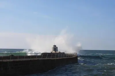 a lighthouse in the ocean with a large wave at Inner Harbour Cottage in Portreath