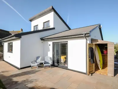 a white house with a patio with two chairs and a surfboard at Zephyr in Helston